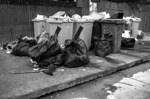 A row of seven large wheeled rubbish bins aligned on a concrete pavement, with some lids open and items such as paper, cardboard, and plastic waste visible inside. Several black plastic rubbish bags are placed on the ground next to the bins, some appearing to be full and tied at the top, with loose waste and paper debris scattered nearby. The background features a wooden fence, and the bins are positioned on a slightly elevated area of the pavement, which shows signs of wear and minor cracks. The scene appears to be part of a waste collection or rubbish removal service in a residential or commercial area, consistent with the services offered by Garden Clearance Hackbridge near the postcode matching the area of London.