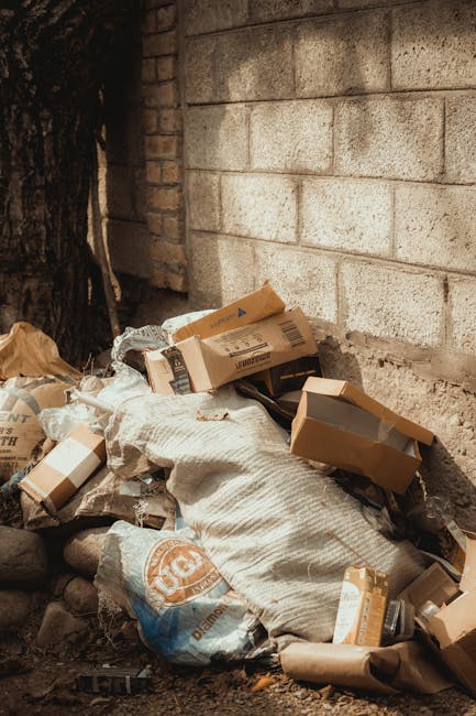 A weathered red metal skip with vertical ridges and a slightly rusted surface is positioned against a grey concrete wall on a paved sidewalk. Next to the skip, several clear plastic bags filled with m