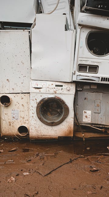 The image features a stack of discarded white household appliances placed outdoors against a background of a muddy ground with scattered leaves and debris. Prominently visible is a front-loading washi